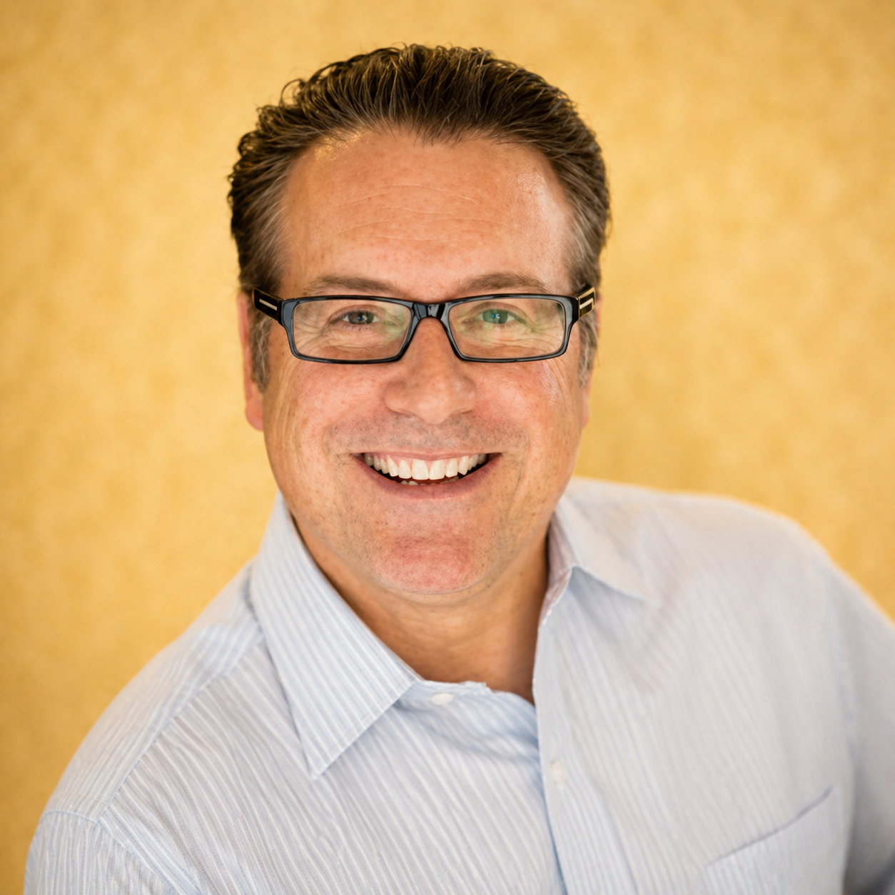 A man wearing glasses and a light blue shirt smiling against a yellow background.