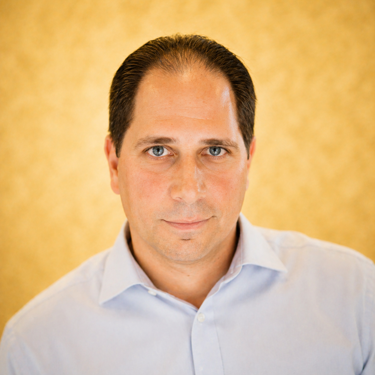 A man in a light blue shirt with a neutral expression against a yellow patterned background.