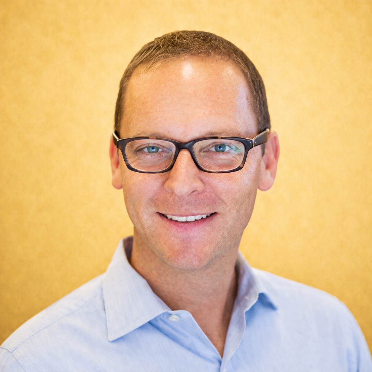A man in glasses smiling against a yellow background.