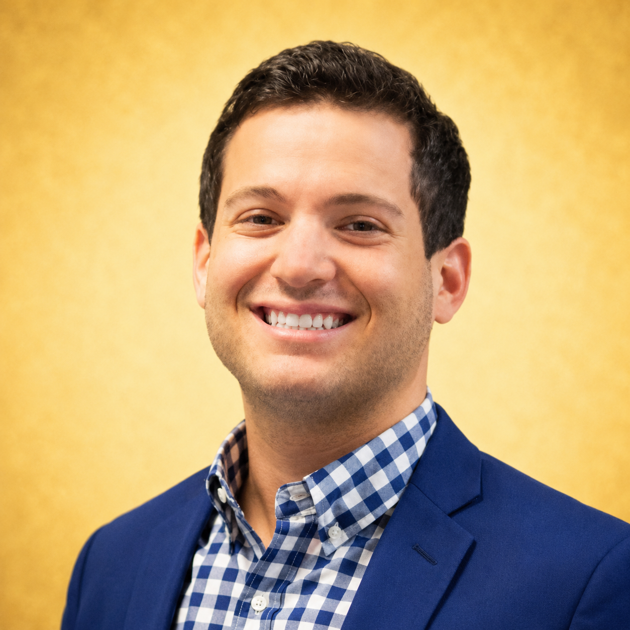 A man in a blue suit and checkered shirt smiling against a yellow background.