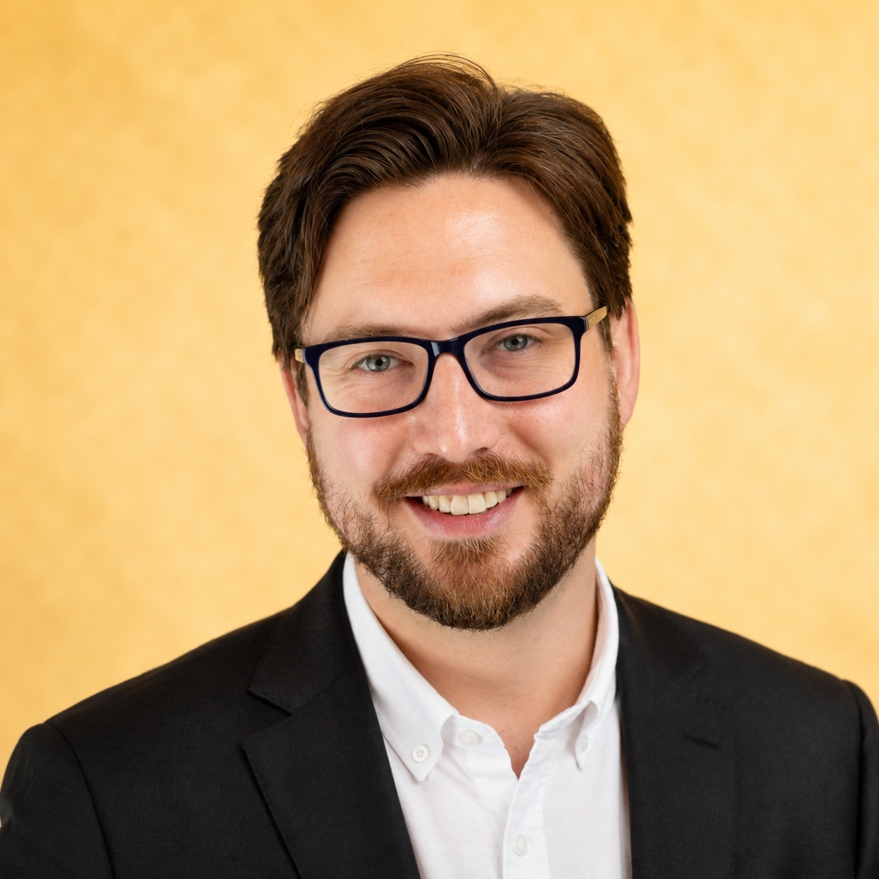 A man with glasses and a beard wearing a suit, smiling against a gold background.