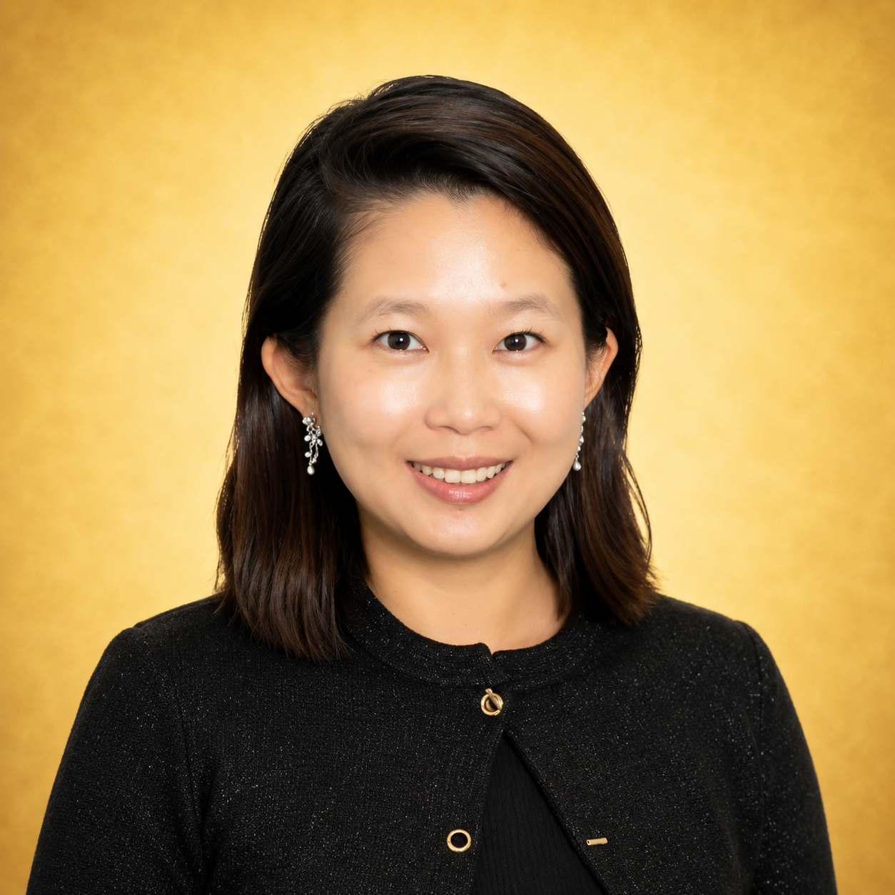 Woman smiling in front of a gold background, wearing a black jacket and earrings.