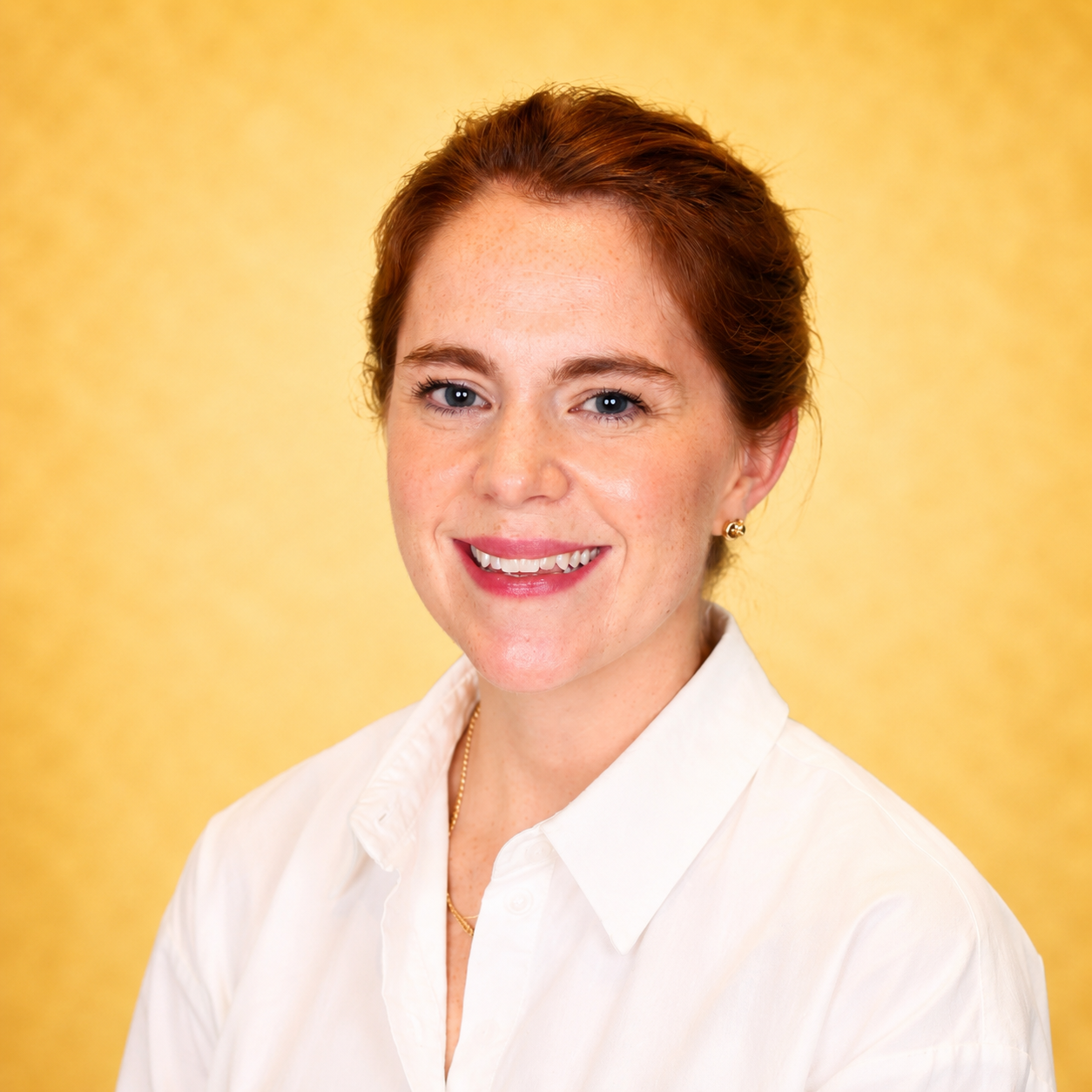 A woman with red hair smiling in a white shirt against a yellow background.