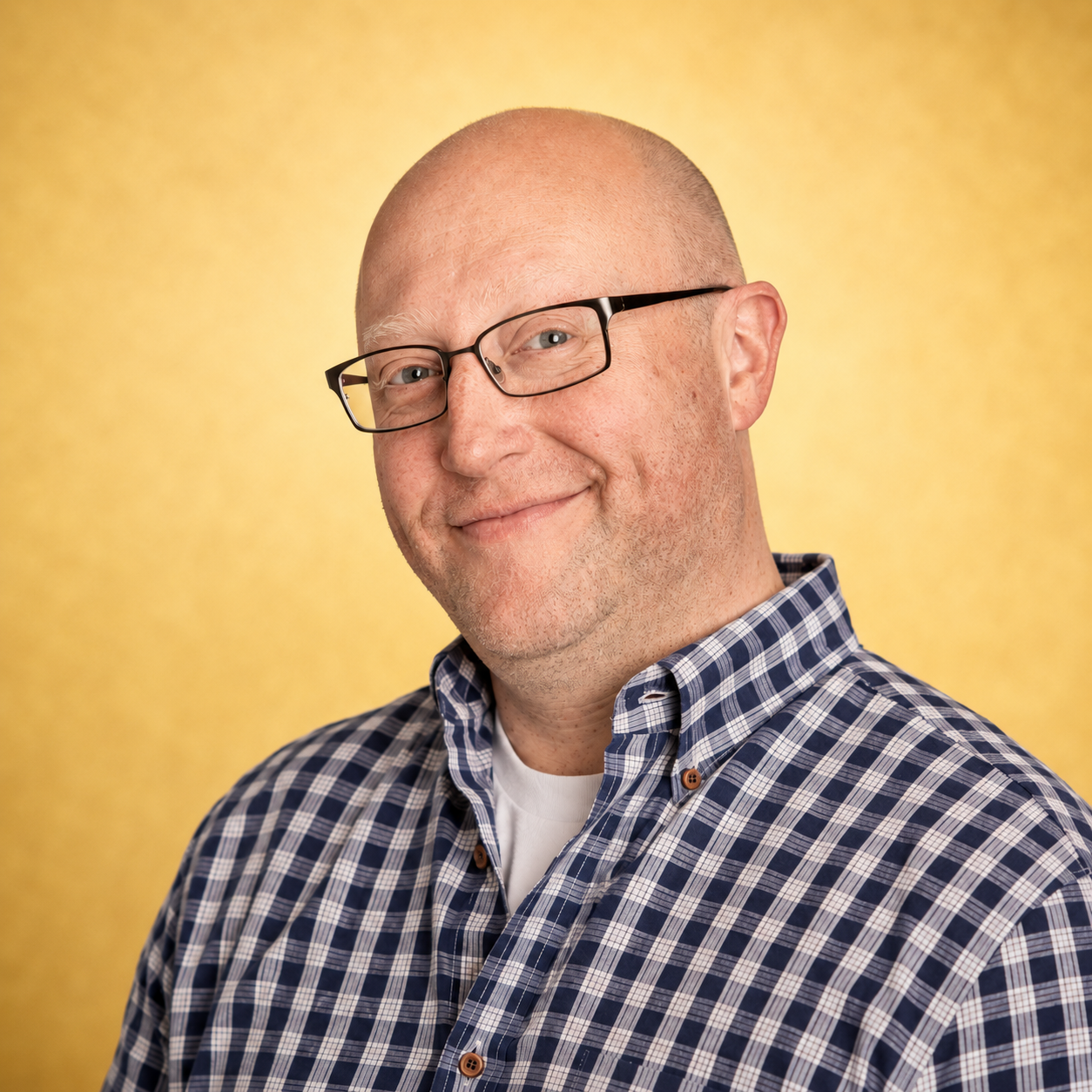 A smiling man in glasses and plaid shirt against a yellow background.