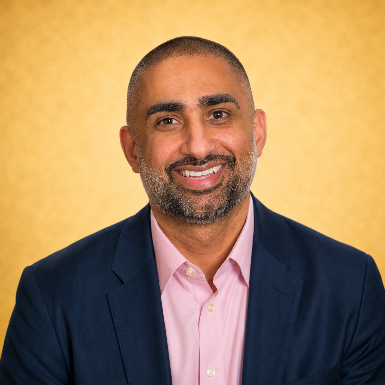 A smiling person in a navy blazer and pink shirt against a yellow background.