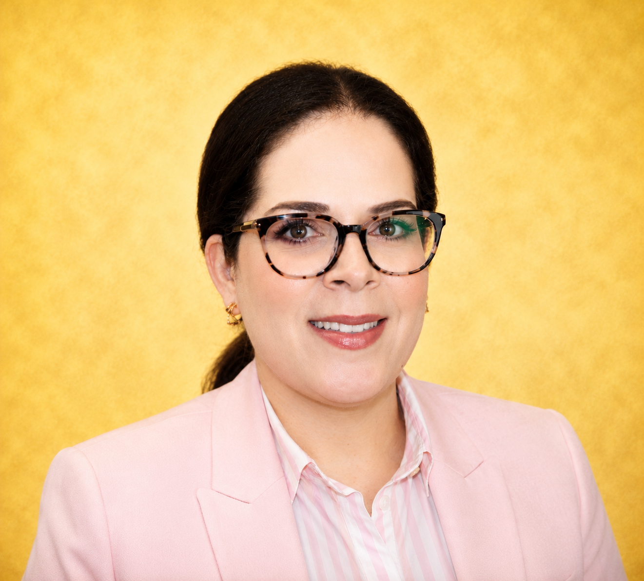 A woman in glasses and a pink blazer smiles against a yellow background.