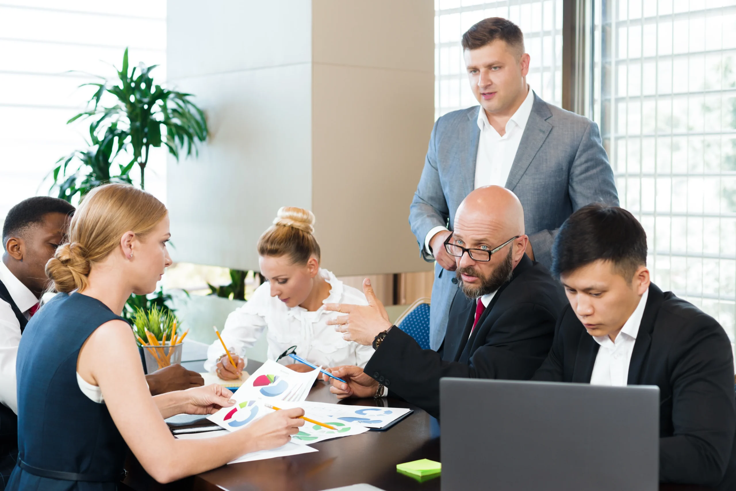 A man standing in a business meeting as team members review charts and discuss data around a conference table