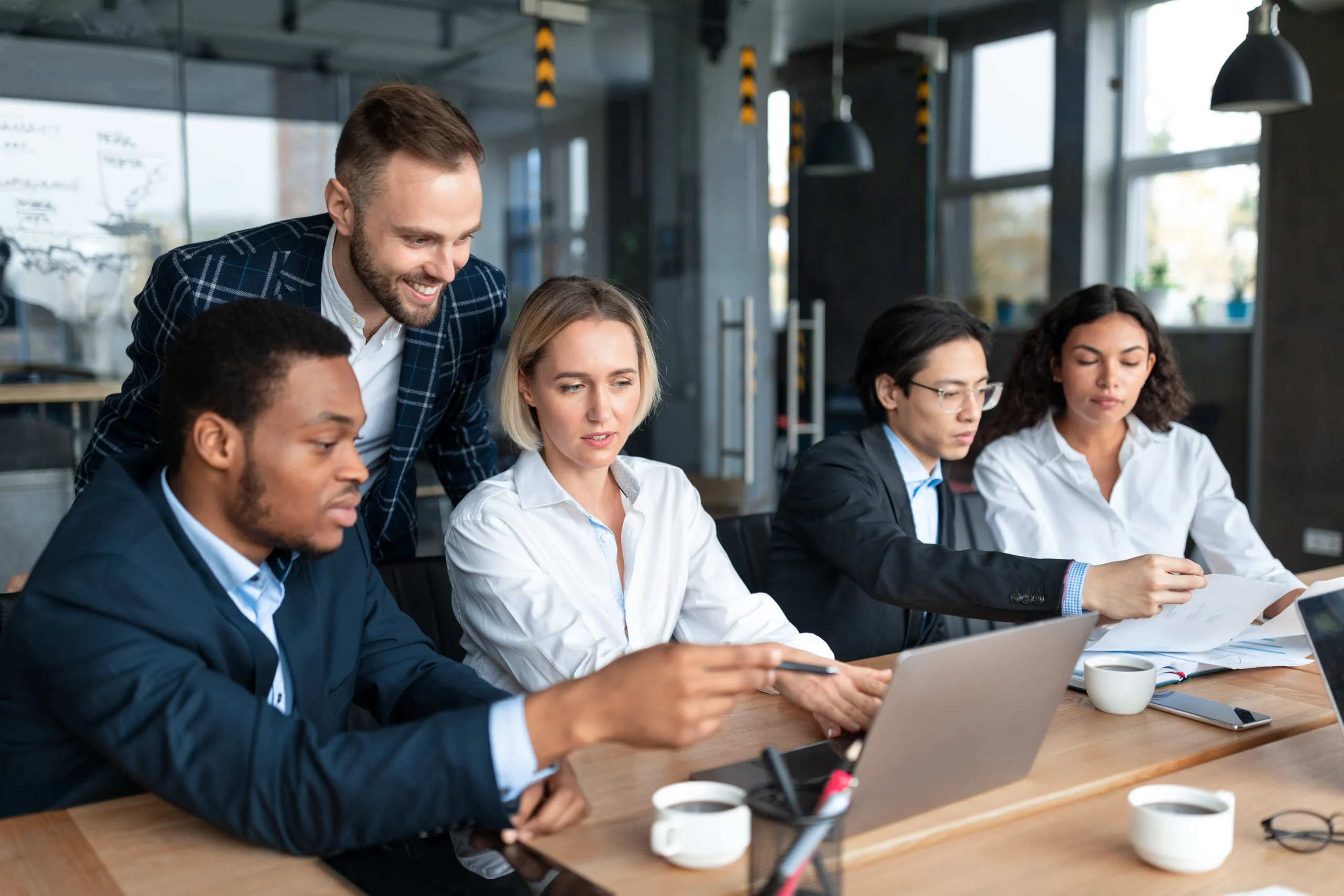 Business team (two women, three men) collaborating over laptop and documents in a modern office.