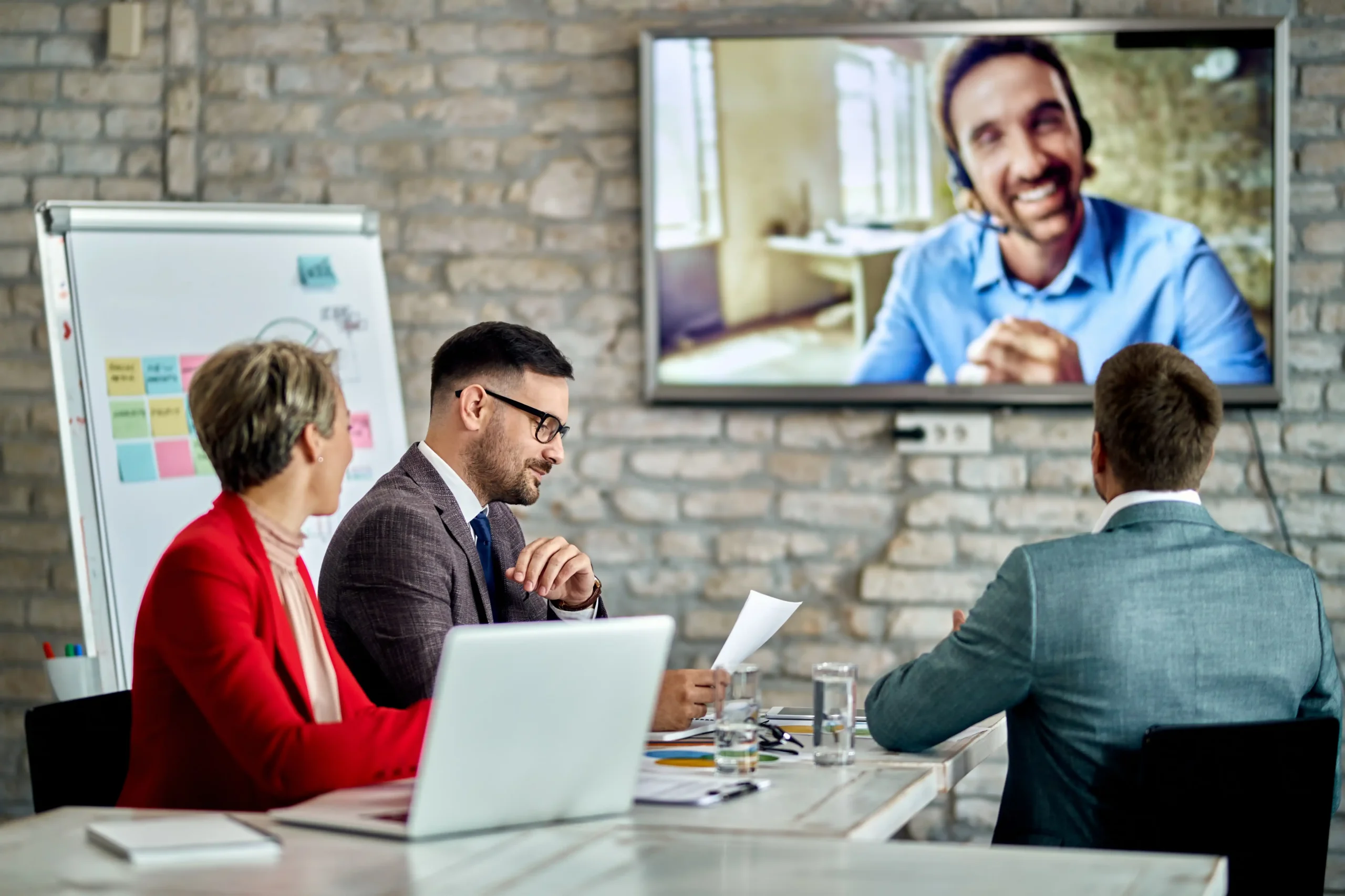 Team in a business meeting with laptops and whiteboard, watching a smiling man on a large video conference screen.