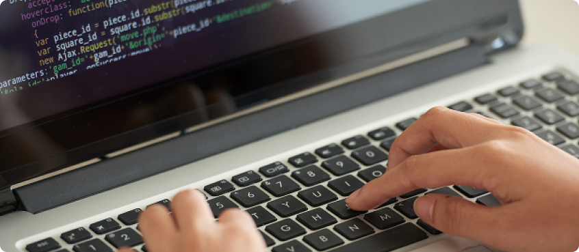 A person typing on a laptop keyboard with programming code visible on the screen