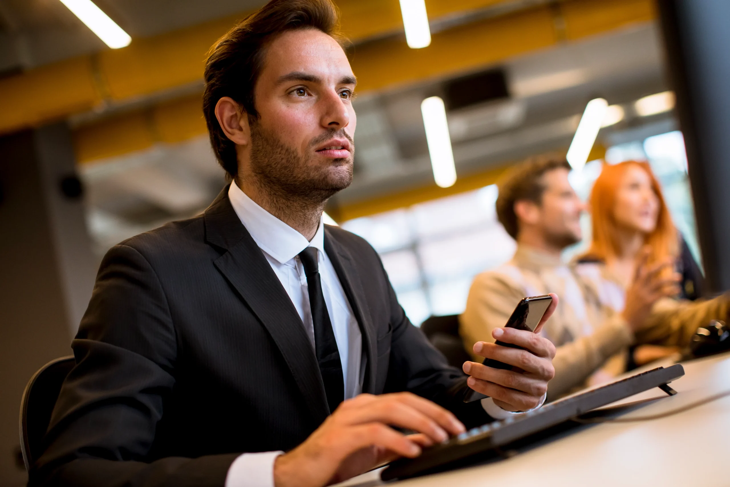 A man in a suit holding a smartphone while typing at a keyboard in a modern open-plan office
