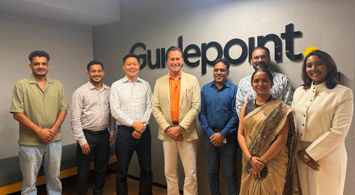 Group of eight professionals smiling in front of Guidepoint office sign, business casual and formal attire.