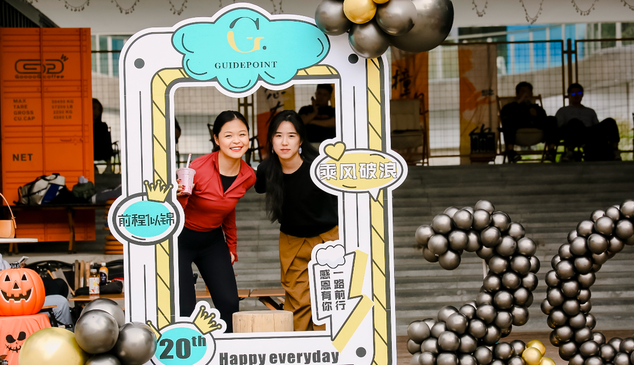 Two women posing inside a colorful Guidepoint photo-frame prop with balloons at an outdoor event, one holding a drink