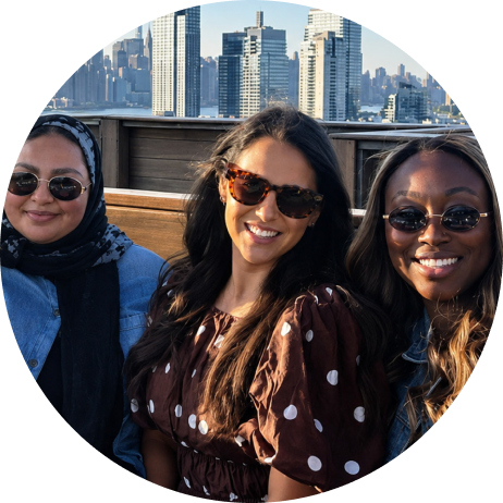 Three smiling women in sunglasses sitting on a rooftop terrace with an urban skyline behind them