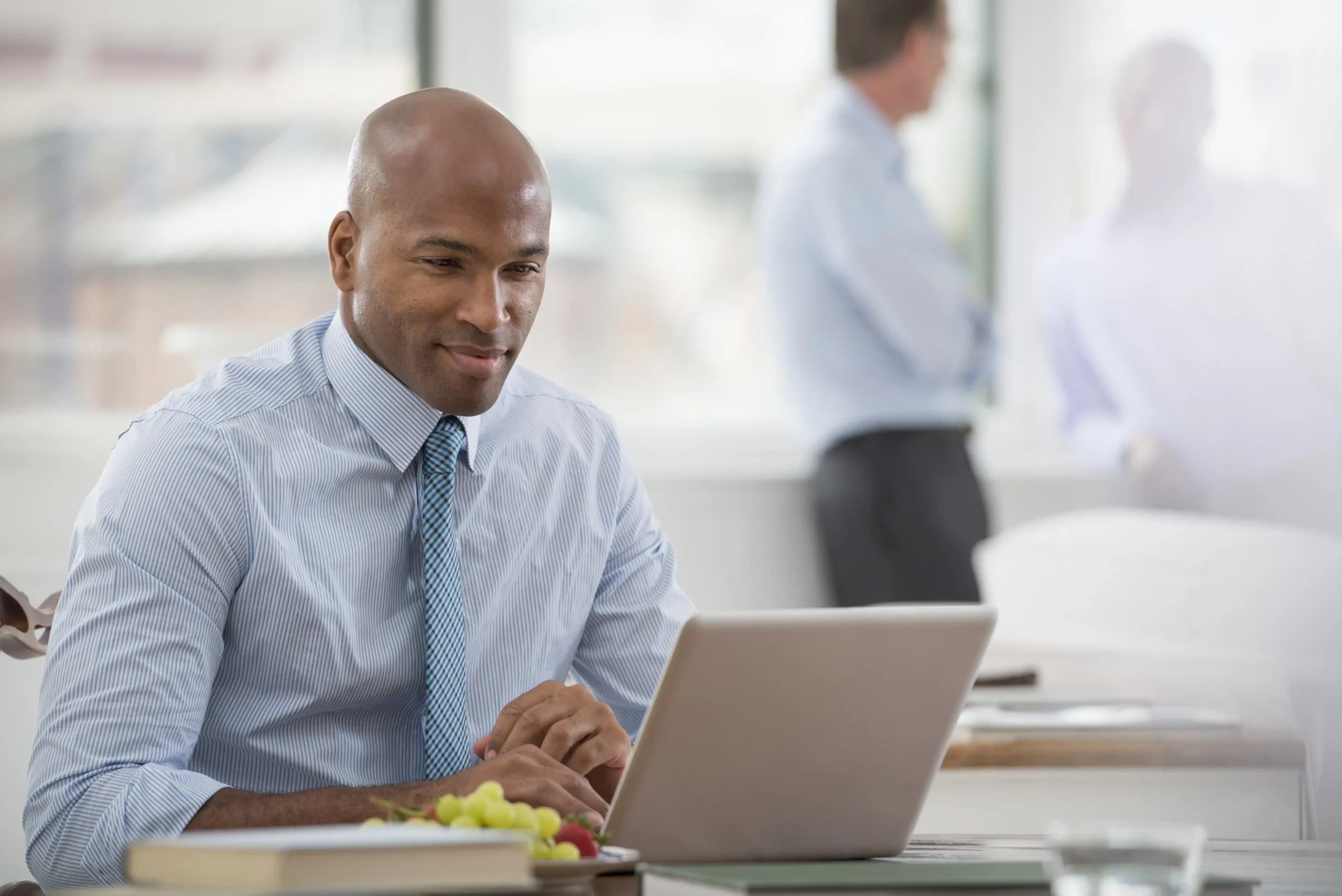 A man in a striped shirt and blue tie working on a laptop at an office desk, blurred colleagues in background
