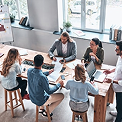 Team meeting: six colleagues collaborating around a wooden table with laptops, papers and coffee in a bright office.