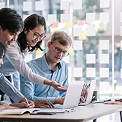 Three colleagues collaborating around a laptop in a bright office, pointing at screen with sticky notes on glass wall.