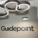Guidepoint logo with yellow dot on gray office wall beneath modern circular LED light fixtures.