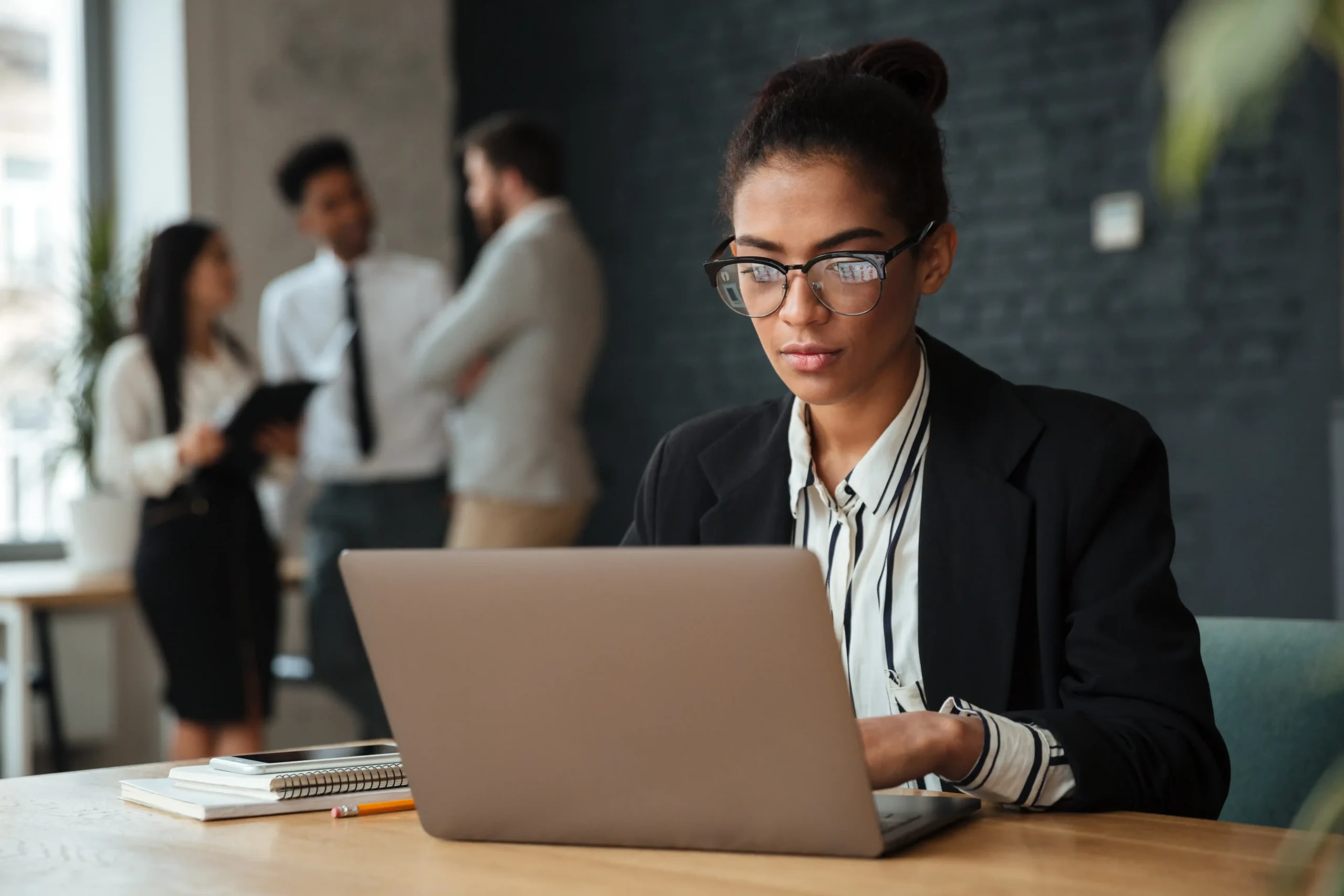 A woman in glasses focused on a laptop at a modern office desk with colleagues talking in the background.